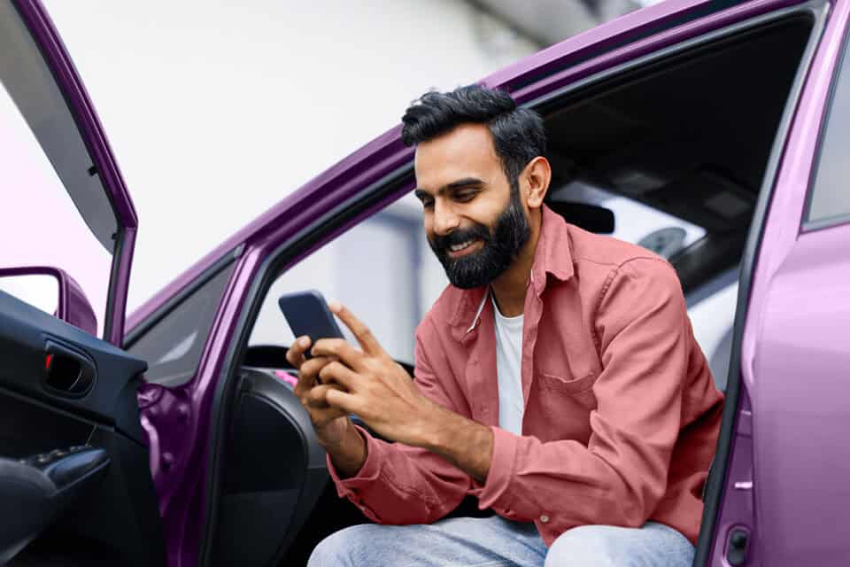 Smiling man using smartphone filling out an auto lease quote request while sitting in a purple car.
