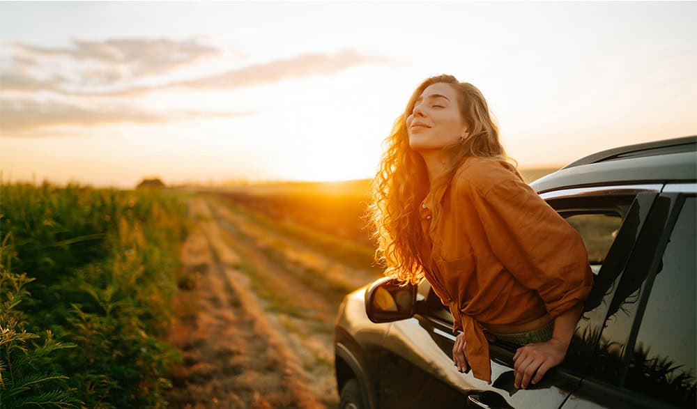 Woman leaning out of car window at sunset, enjoying the view in a rural setting in her older auto purchase.