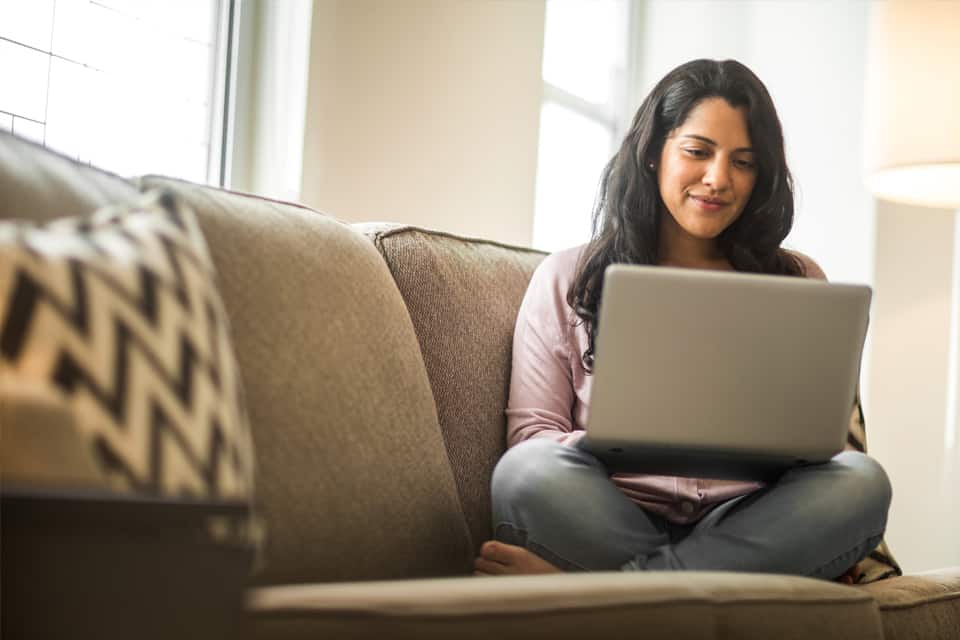 Una mujer sentada en un sofá mientras trabaja en su computadora portátil usando nuestras calculadoras financieras de pagos, como la calculadora de hipotecas.