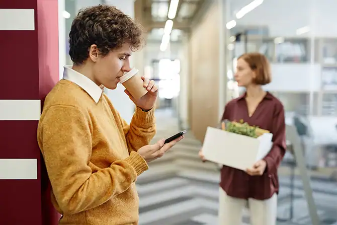 Young man drinking coffee and using smartphone in office corridor to read up on fake job scams.