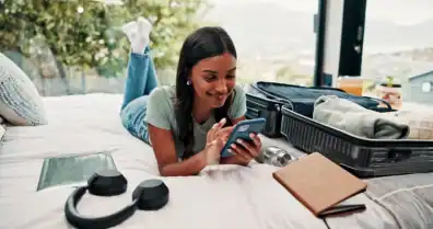 Young happy woman relaxing on bed with phone, travel essentials, and suitcase nearby.