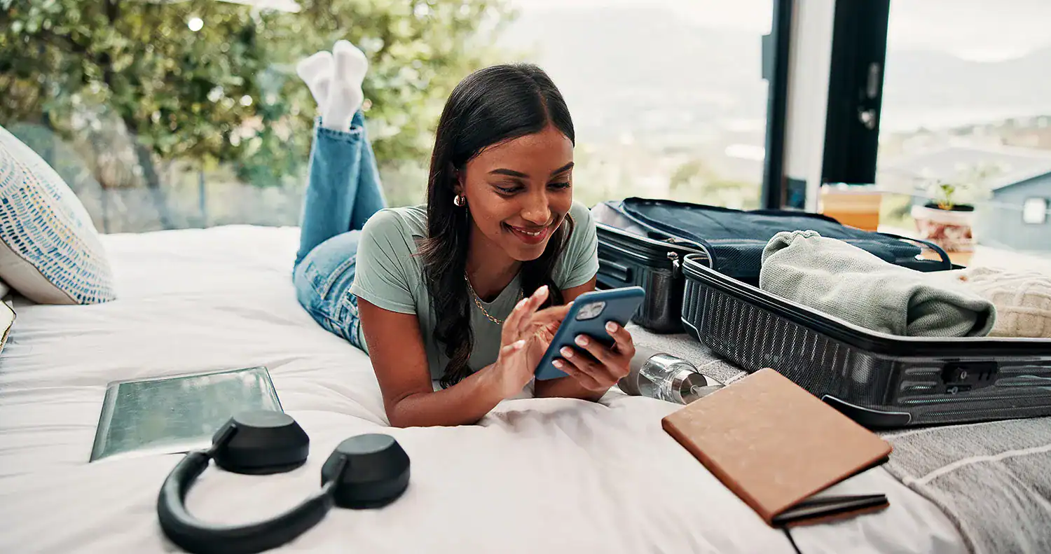Young happy woman relaxing on bed with phone, travel essentials, and suitcase nearby.
