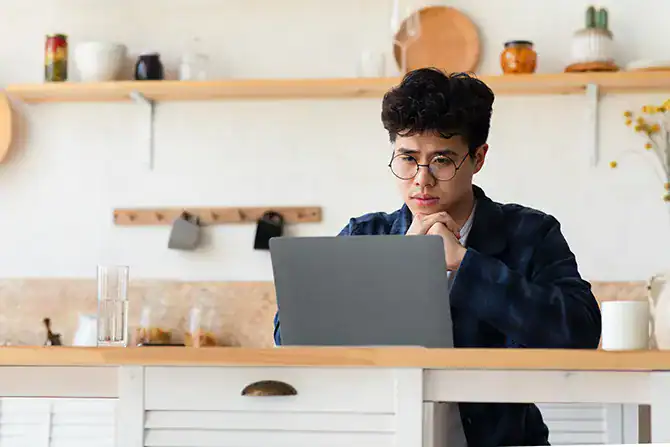 Young man working on a laptop learning about wire and cryptocurrency scams while sitting comfortably at a kitchen table.