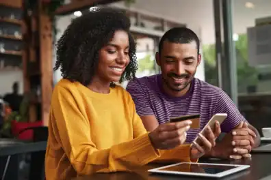 Happy diverse couple using smartphone at cafe, promoting financial education.