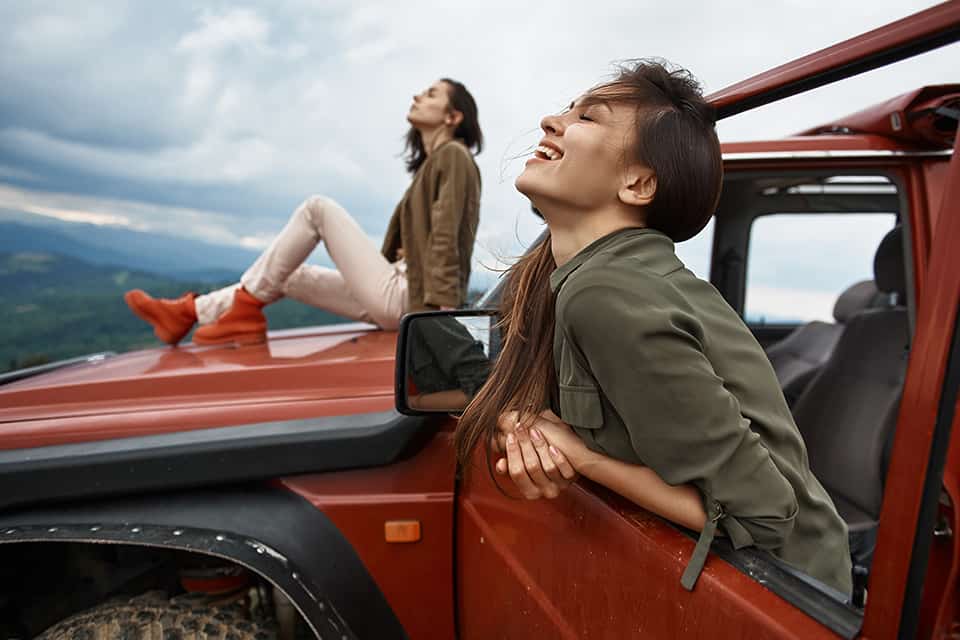 Una mujer se sienta en el capó de un auto mientras otra sonríe.