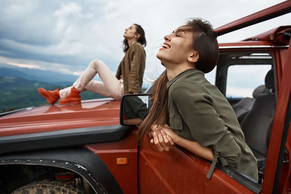 One woman sits on the hood of a car while another smiles