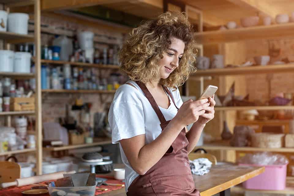 A woman stands in a pottery studio and looks at her mobile phone