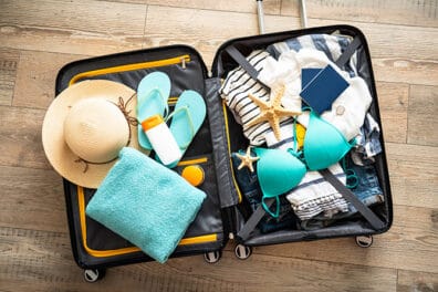 Open suitcase with summer travel essentials including hat, towel, flip-flops, and swimwear.